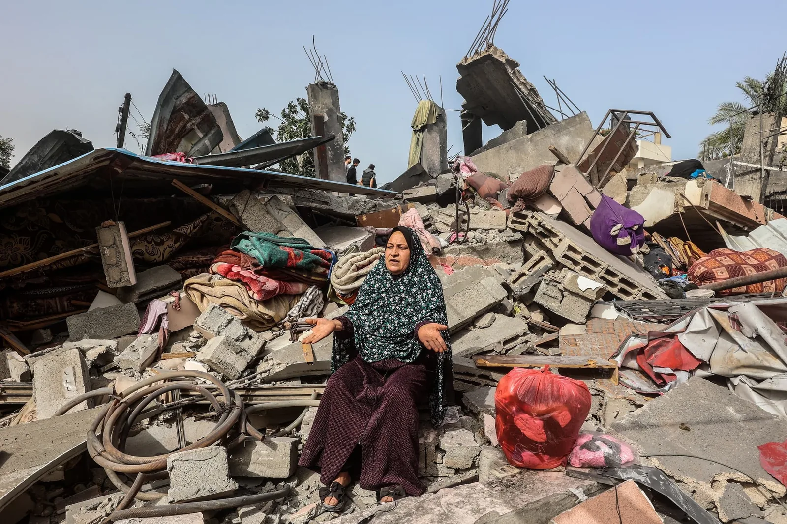 A woman sits among concrete rubble and household items in a damaged urban area.