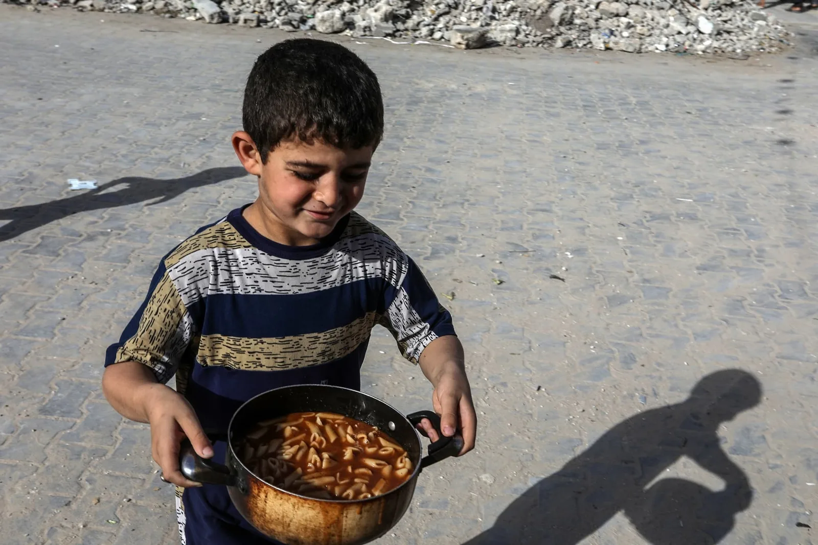 A child carries a large cooking pot filled with food across a paved outdoor area.