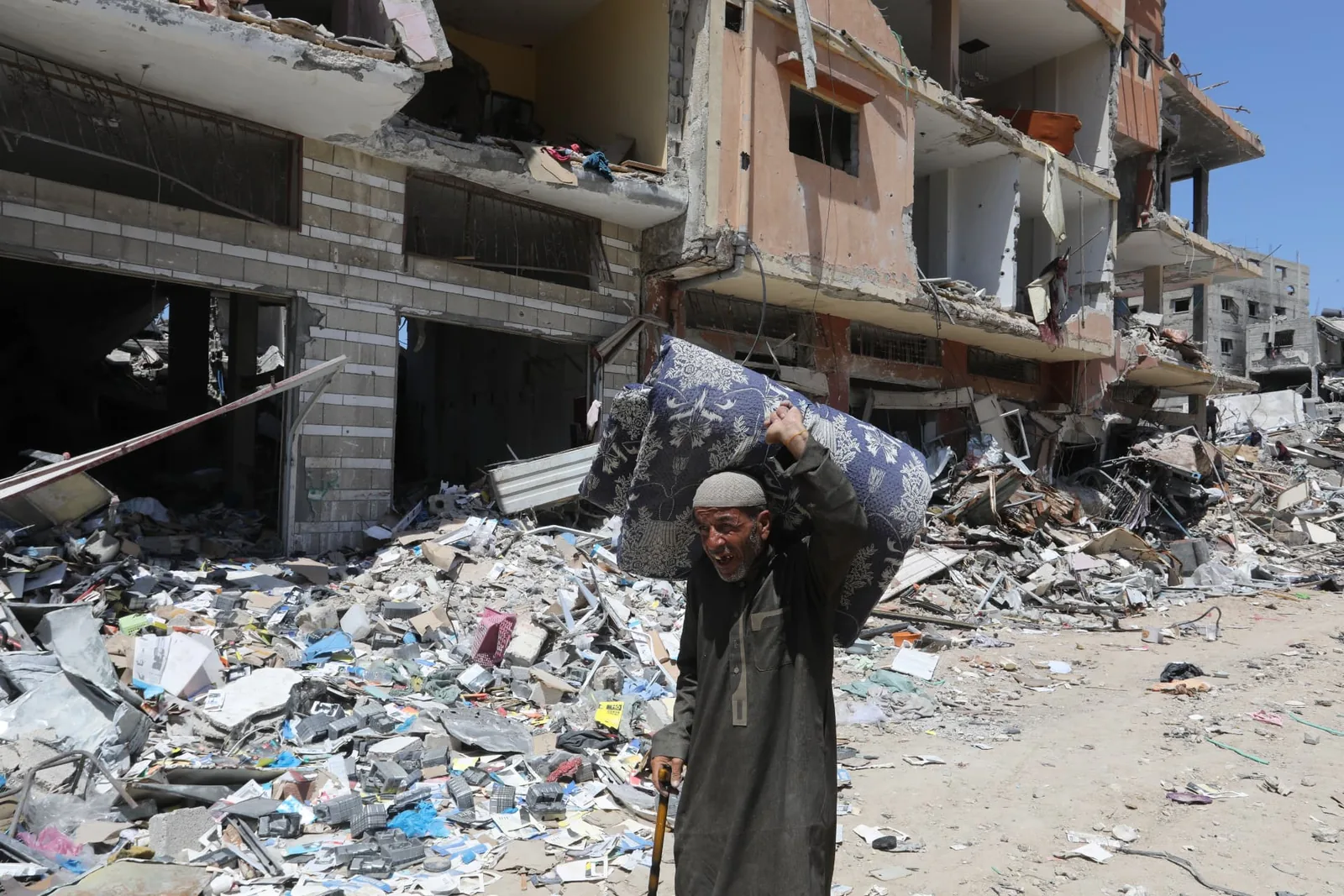 An older adult carries a large bedding bundle through an area of damaged buildings and rubble.