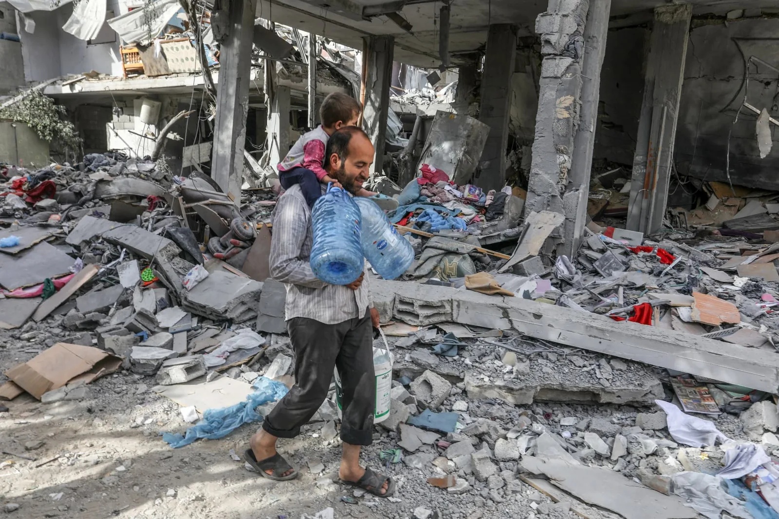 An adult carries water containers and a child through a heavily damaged area.