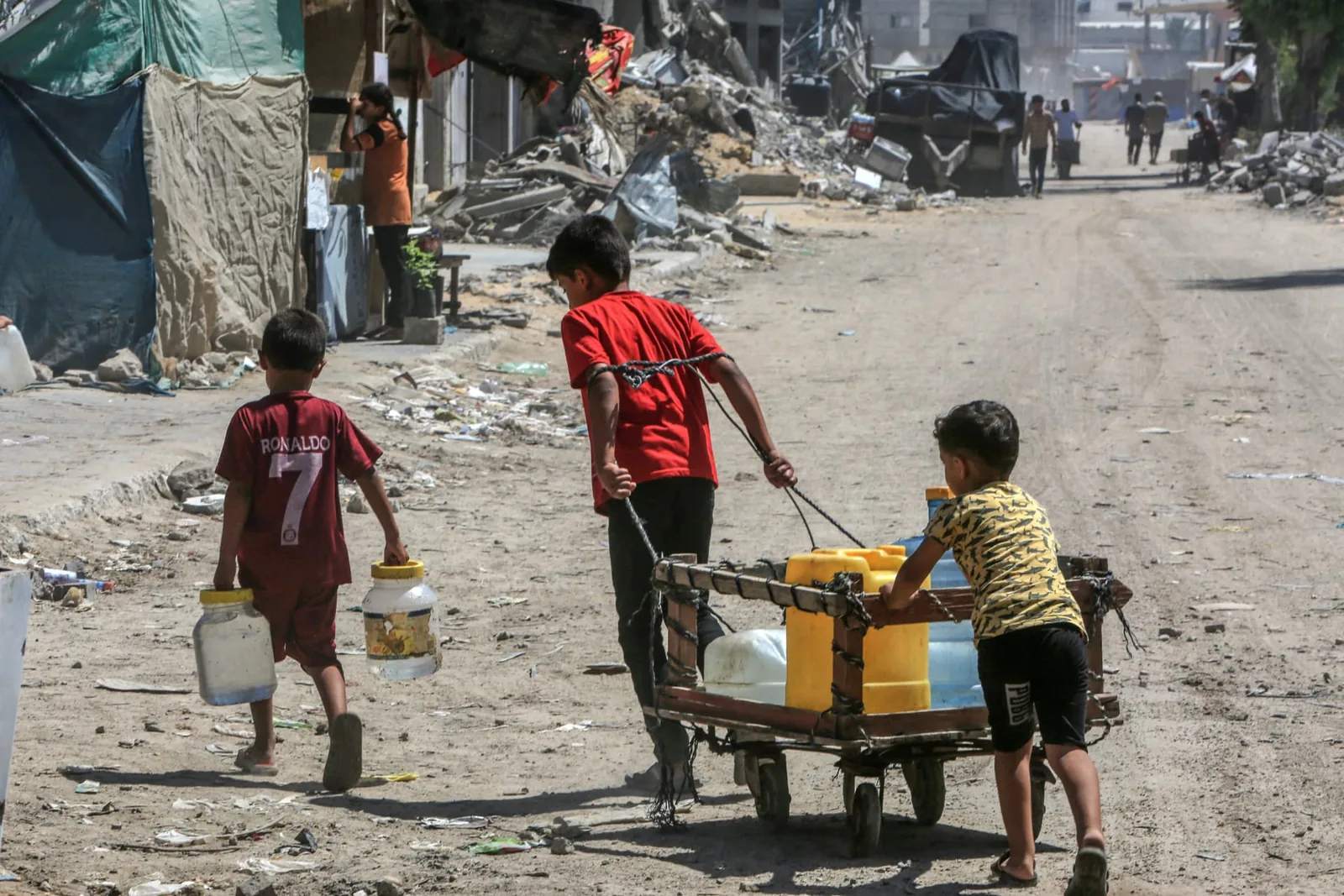 Three children move yellow water containers on a small cart through a damaged neighborhood.